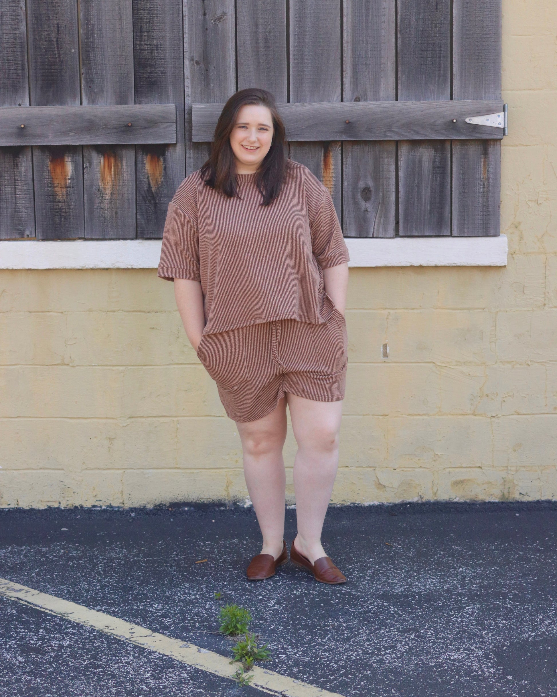 Woman in brown outfit standing in front of a wooden fence and yellow wall.