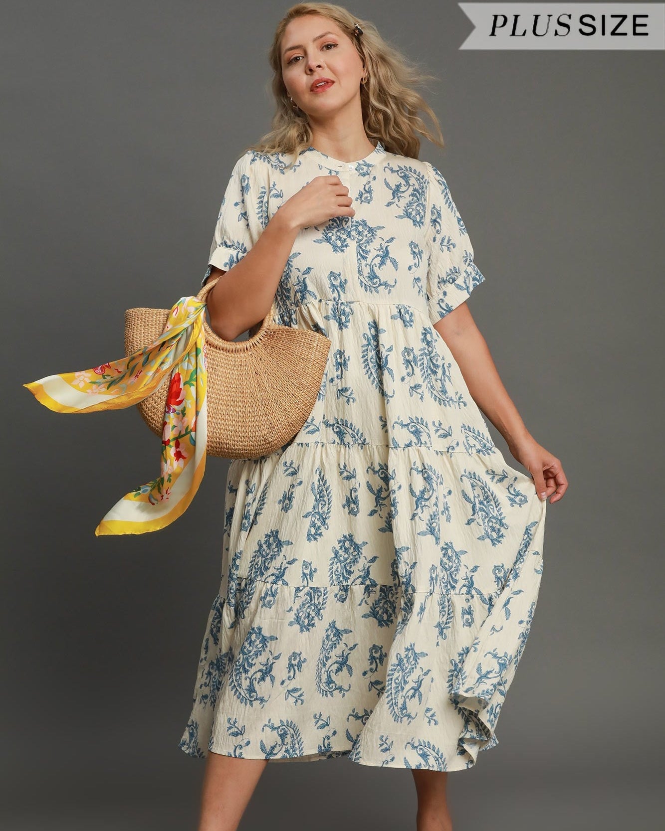 Woman wearing a floral dress holding a bag and scarf against a gray background