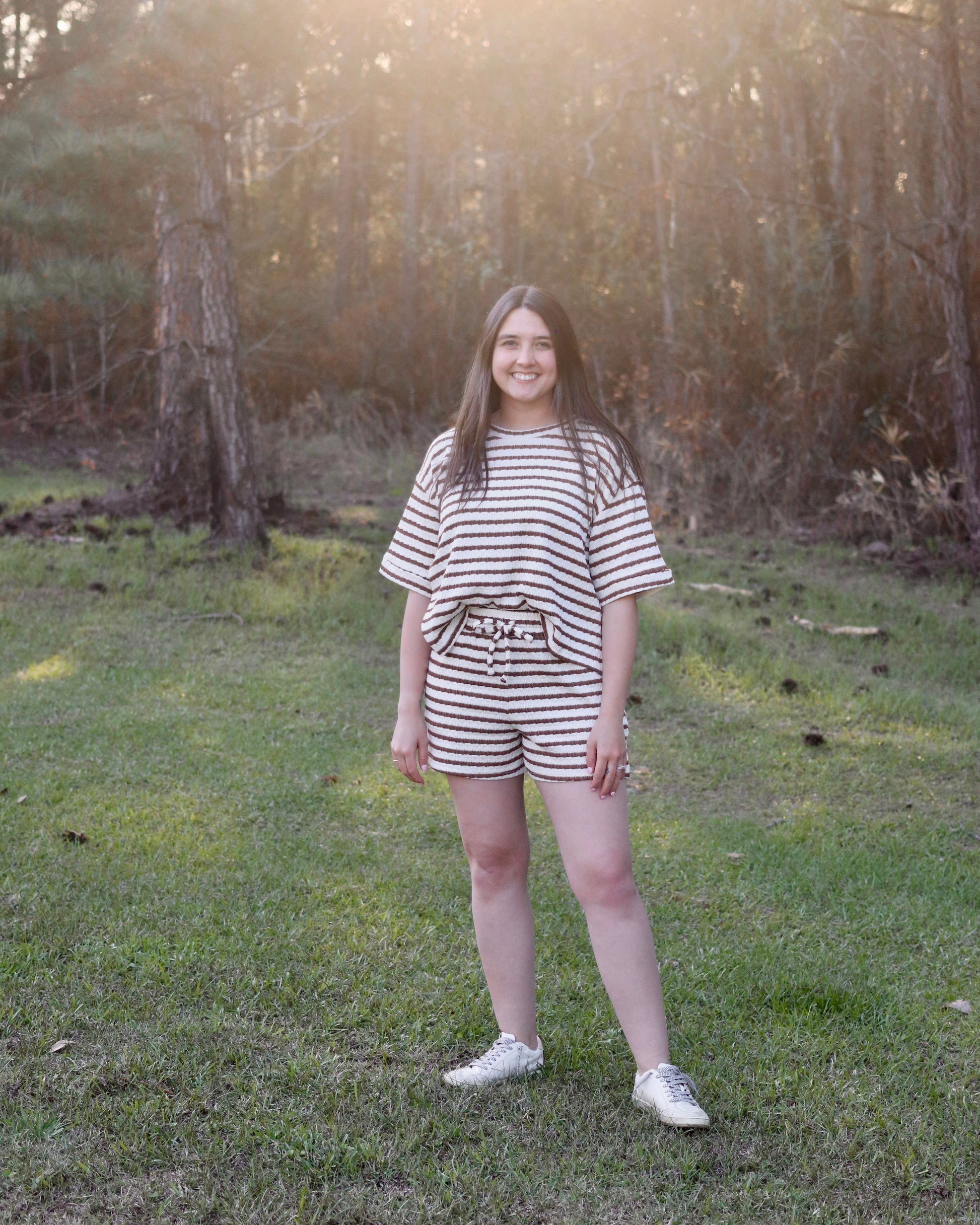 Person wearing a striped outfit standing in a forest with sunlight filtering through the trees.
