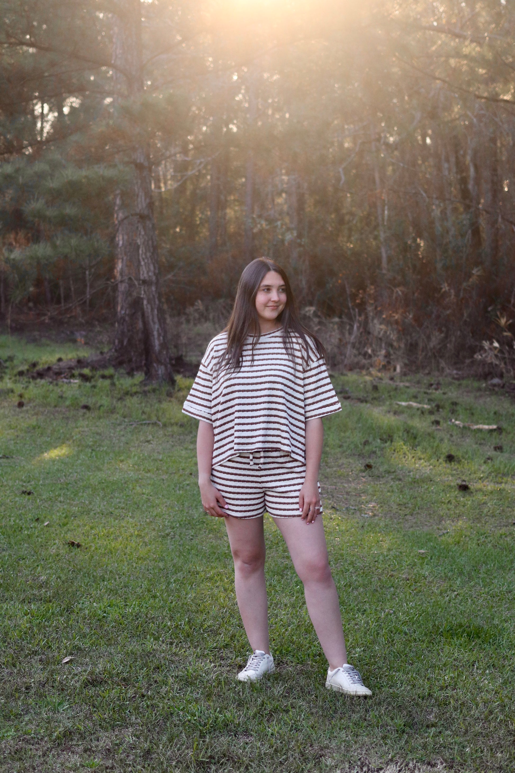 Person wearing a striped outfit standing in a forest with sunlight filtering through the trees.