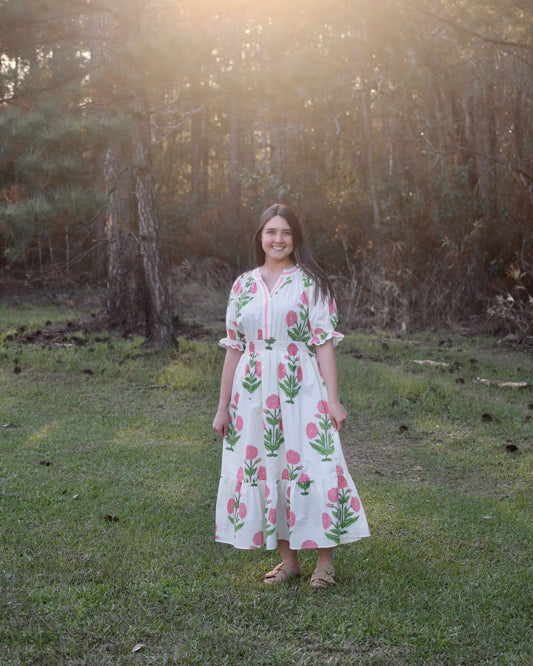 Woman in a floral dress standing in a grassy field with sunlight filtering through trees.