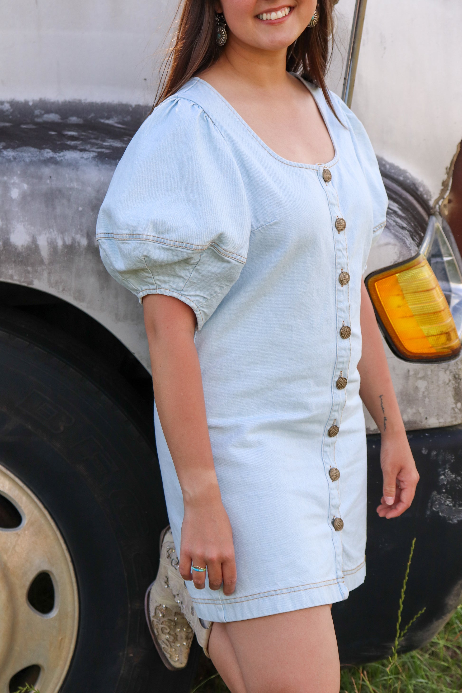 Woman wearing a light blue denim dress standing next to a vehicle.