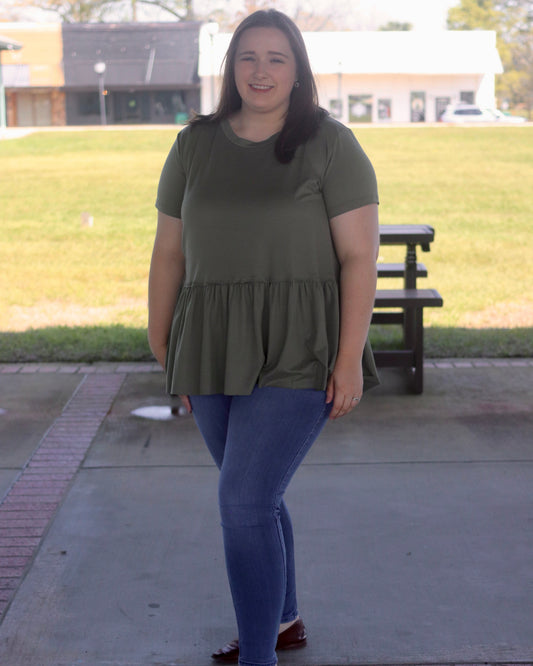 Woman wearing a green top and blue jeans standing on a sidewalk with grass and a building in the background.