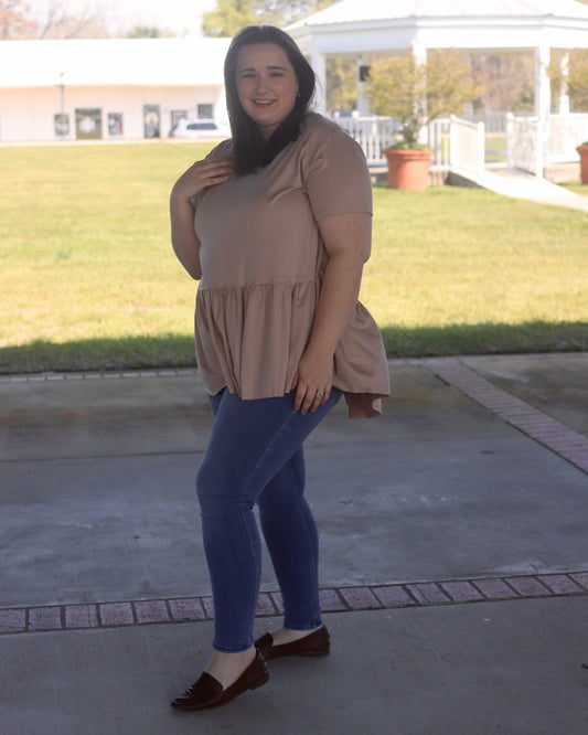 Woman wearing a beige top and blue jeans standing on a concrete patio with grass and a gazebo in the background.