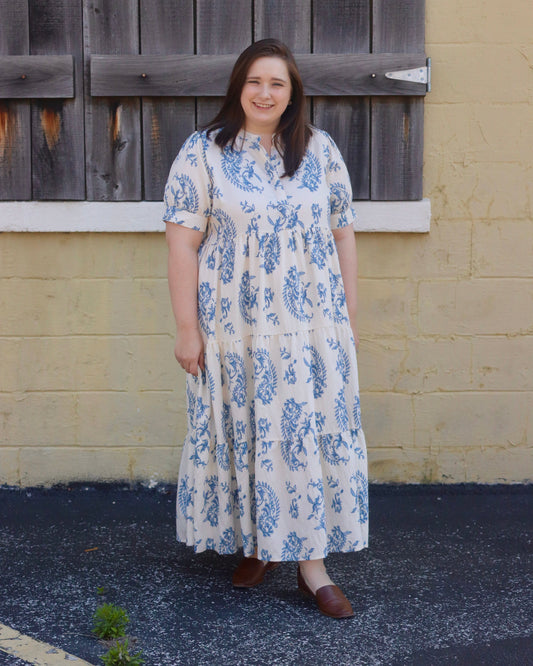 Woman wearing a floral dress standing in front of a wooden shutter and yellow wall.